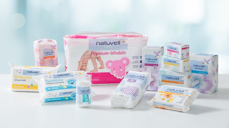 Mehrere naturwell-Produkte wie Feuchttücher, Windeln und Einlagen stehen auf einem weißen Tisch vor unscharfem, blauem Hintergrund.