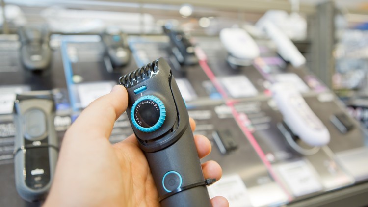 Eine Hand hält einen schwarzen Barttrimmer mit blauem Einstellrad vor einem Regal mit weiteren Rasierern.