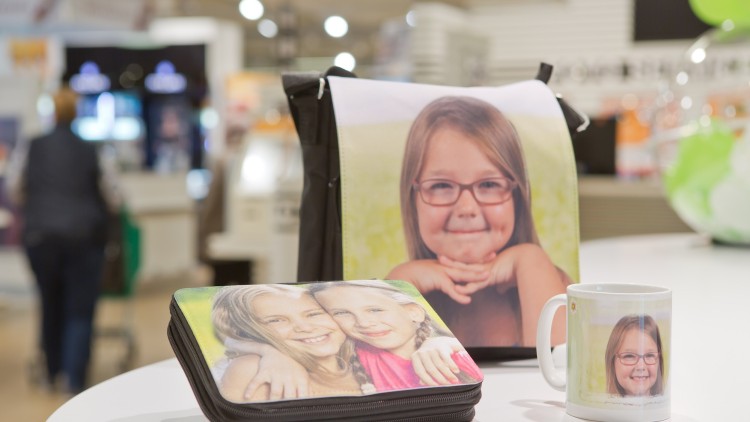 Tasche, Fotobuch und Tasse mit einem individuell bedruckten Motiv stehen auf einem weißen Tisch in einer Geschäftsumgebung.