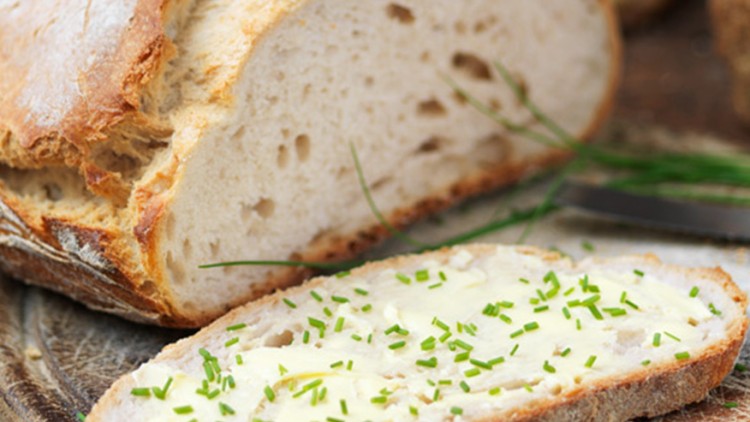 Ein aufgeschnittenes Brot mit Butter und Schnittlauch liegt auf einem Holzbrett, daneben ein Messer und weitere Backwaren, Text: Meisterbäckerei, Hauseigene Herstellung, GLOBUS.