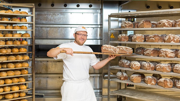 Bäcker holt mit einem langen Holzschieber frisch gebackenes Brot aus einem Metallregal in einer Backstube.