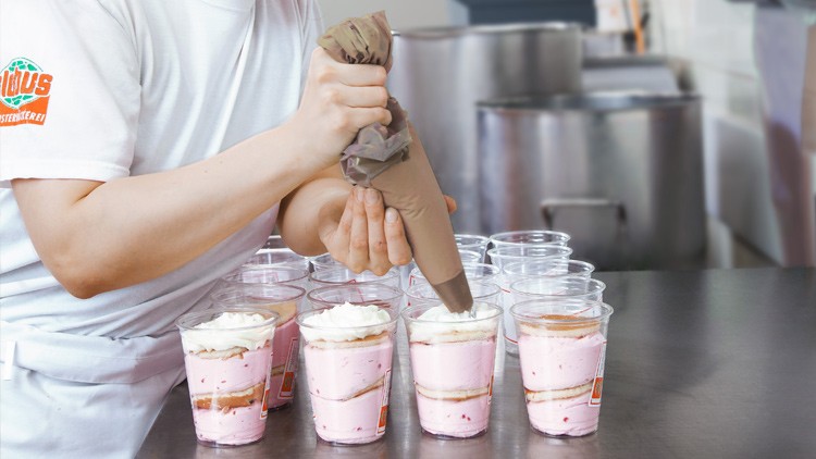 Eine Person füllt mit einem Spritzbeutel weiße Creme auf rosafarbenes Dessert in durchsichtigen Bechern mit GLOBUS-Logo.
