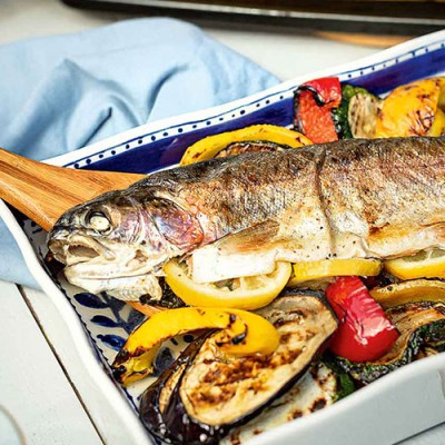Ein ganzer gegrillter Fisch liegt auf gebratenem Gemüse wie Zucchini, Paprika und Aubergine in einer blauen Auflaufform.