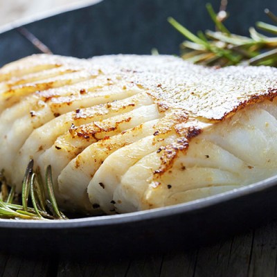 Gebratener Fisch mit goldbrauner Kruste und angeröstetem Rosmarin in einer dunklen Pfanne auf Holzuntergrund.