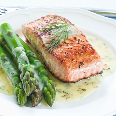 Gegrilltes Lachsfilet mit frischem Dill auf heller Soße, daneben grüne Spargelstangen auf einem weißen Teller.