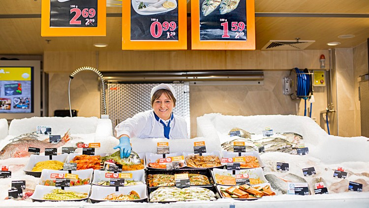 Frischetheke mit Fisch und Meeresfrüchten, Preisschildern und weiß bekleidetem Personal vor einem gekachelten Hintergrund.
