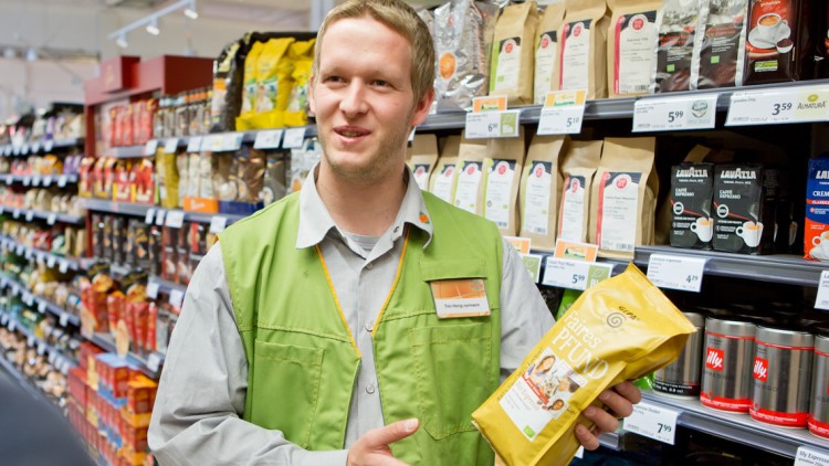 Eine Person in GLOBUS-Arbeitskleidung hält eine gelbe Kaffeeverpackung vor einem Regal mit verschiedenen Kaffeesorten.