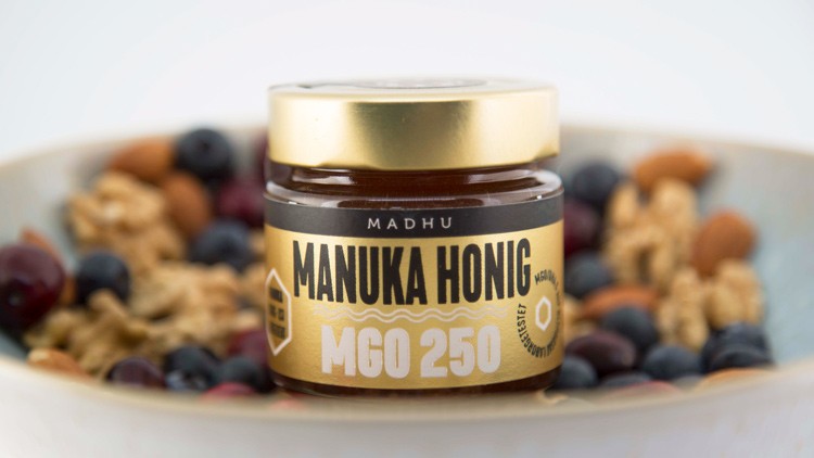 Ein Glas „Madhu Manuka Honig MGO 250“ mit goldfarbenem Deckel steht vor einer Schale mit Nüssen, Beeren und Müsli.