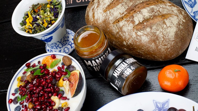 Ein Laib Brot, ein Glas Manuka-Honig, ein Müsli mit Früchten, ein Teebecher, eine Mandarine und getrocknete Kräuter auf schwarzem Tisch.