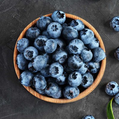 Holzschale mit frischen Blaubeeren, daneben verstreute Blaubeeren und grüne Blätter auf dunklem Untergrund.