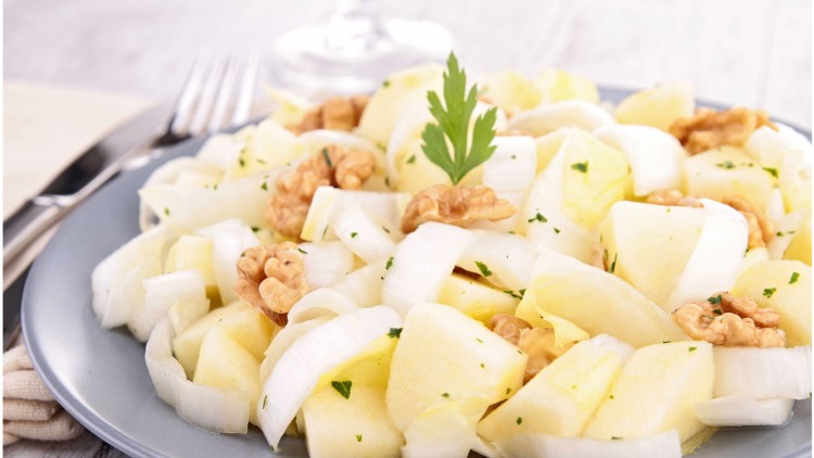Chicorée-Salat mit Apfelstücken, Walnüssen und Petersilie auf einem grauen Teller, im Hintergrund Besteck und ein Glas.