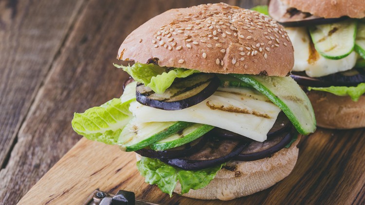 Veggie-Burger mit Sesambrötchen, gegrillter Zucchini, Aubergine, Halloumi und frischem Salat auf Holzbrett.