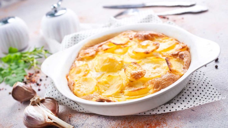 Kartoffelgratin mit goldbrauner Kruste in einer weißen Auflaufform, daneben frischer Knoblauch sowie Kräuter auf einem Tuch mit Punkten.