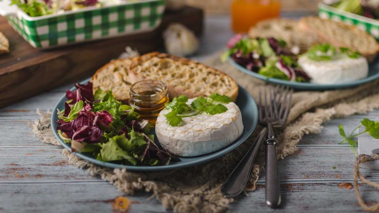 Gebackener Camembert mit frischen Salatblättern, Kräutern, Honigglas und Brot auf einem blauen Teller, Gabel und Messer daneben.