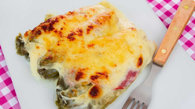 Überbackene Endivien mit Käse, Schinken und cremiger Soße auf einem weißen Teller mit einer Gabel und rosa Karomuster-Serviette.