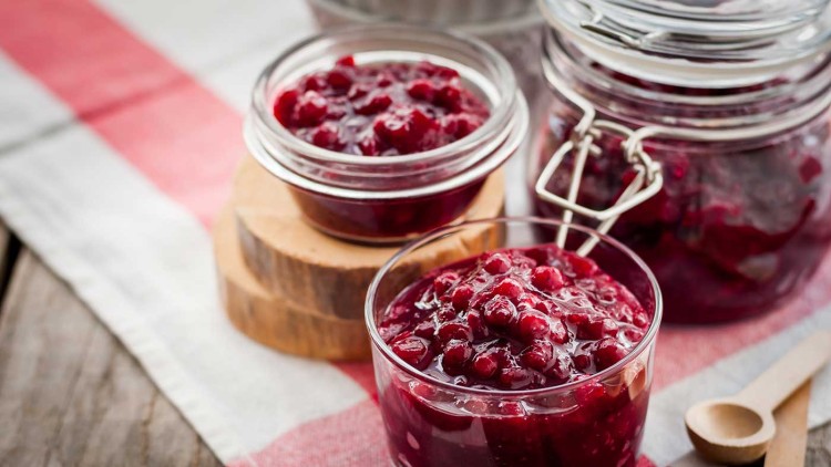 Mehrere Glasbehälter mit rotem Preiselbeermus, Holzlöffel und ein rot-weißes Tuch auf einem Holzuntergrund.