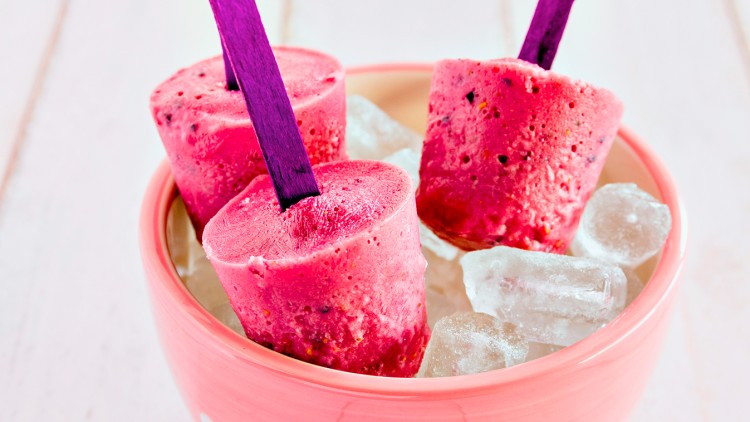 Drei pinke Fruchteislutscher mit lilafarbenen Stielen stehen in einer rosa Schüssel auf Eiswürfeln.