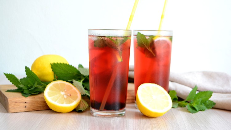 Zwei Gläser mit rotem Eistee, Zitronenscheiben, gelben Trinkhalmen und frischer Minze, daneben ganze und halbe Zitronen.