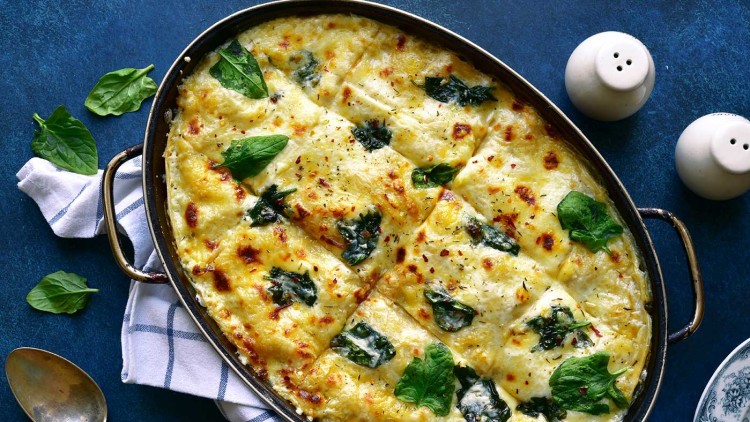 Überbackene Lasagne mit goldbrauner Kruste, frischen Spinatblättern, gewürztem Käse und einem Löffel auf weiß-blauem Tuch.
