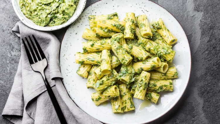 Ein Teller mit Rigatoni und grüner Pesto-Soße, daneben eine Schüssel Pesto, graues Stofftuch und schwarze Gabel.