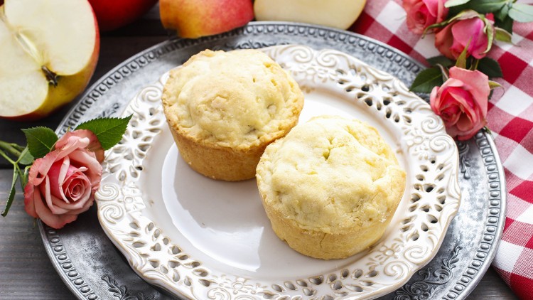 Zwei helle Muffins auf einem verzierten weißen Teller, umgeben von roten Äpfeln, Rosen und einem rot-weiß karierten Tuch.