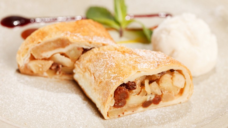 Auf einem Teller liegen zwei Stücke Apfelstrudel mit Puderzucker, ein Klecks Sahne, Minzblätter und eine rote Soßenlinie.