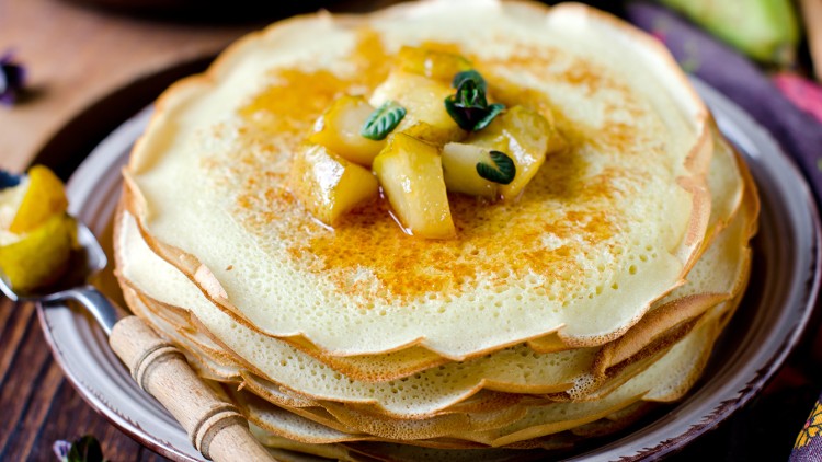 Ein Stapel heller Crêpes auf einem Teller, garniert mit karamellisierten Apfelstücken und kleinen grünen Blättern.