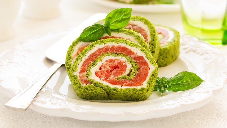 Grüne Spinatrolle mit Schichten aus Frischkäse und rohem Lachs, dekoriert mit frischen Basilikumblättern auf weißem Teller.