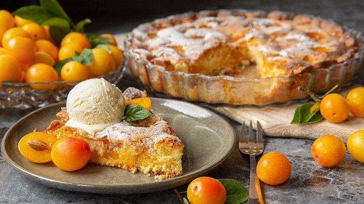 Ein Stück Streuselkuchen mit gelben Früchten, einer Kugel Vanilleeis und Minzblatt auf Teller, daneben Gabel und frische Mirabellen.