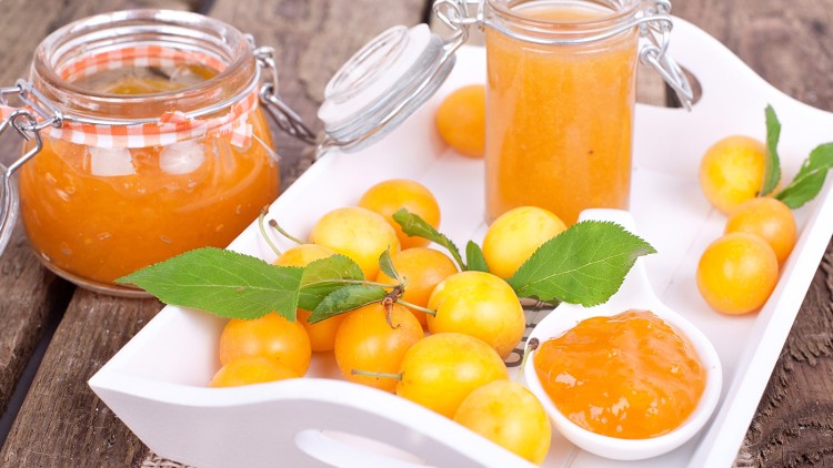 Gelbe Mirabellen mit Blättern liegen auf einem weißen Tablett neben Gläsern und einer Schale mit orangefarbener Marmelade.
