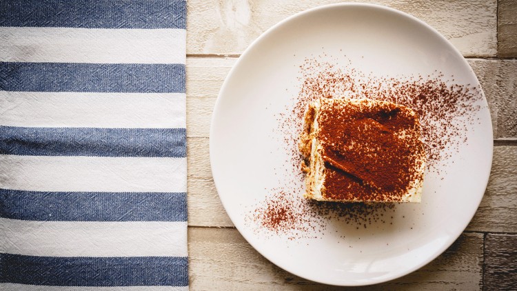 Ein Stück Tiramisu mit Kakaopulver auf einem weißen Teller neben einer blau-weiß gestreiften Serviette auf Holztisch.