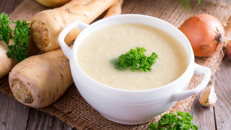 Eine weiße Schale mit cremiger Suppe, dekoriert mit frischer Petersilie, liegt auf einem Holztisch mit Pastinaken, Zwiebel und Apfel.