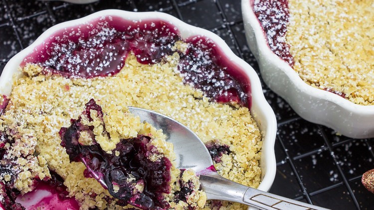 Eine weiße, runde Schale mit warmem Heidelbeer-Crumble, bestreut mit Puderzucker, und einem Löffel, der eine Portion entnimmt.