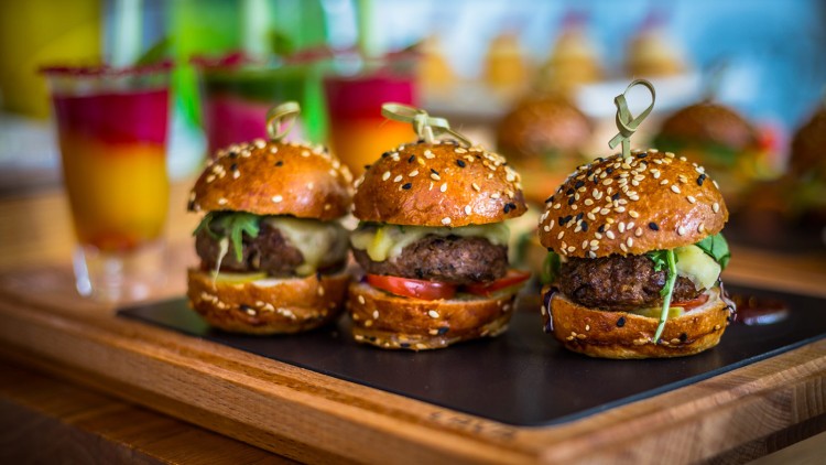 Drei Mini-Burger mit Sesambrötchen, Salat, Tomaten, Käse und Fleischpatty auf einem Tablett, im Hintergrund bunte Getränke.