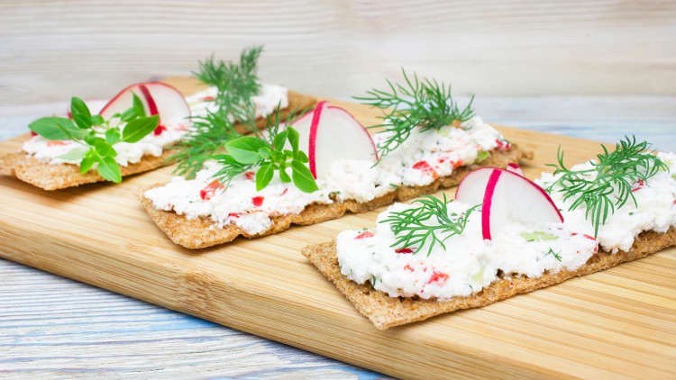 Knäckebrotscheiben mit Frischkäsecreme, Radieschen und frischen Kräutern auf einem Holzbrett.