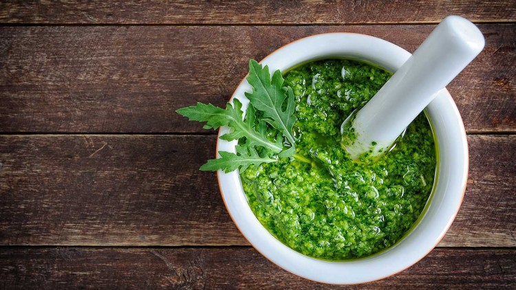Weiße Schale mit grünem Pesto, verziert mit drei Rucolablättern, und einem weißen Mörser auf dunklem Holzuntergrund.