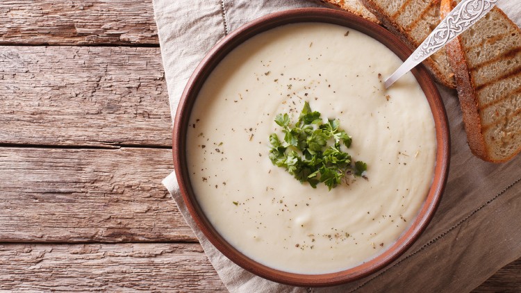 Cremige Suppe in einer braunen Schale, garniert mit frischen Kräutern, daneben geröstete Brotscheiben auf einem Tuch.