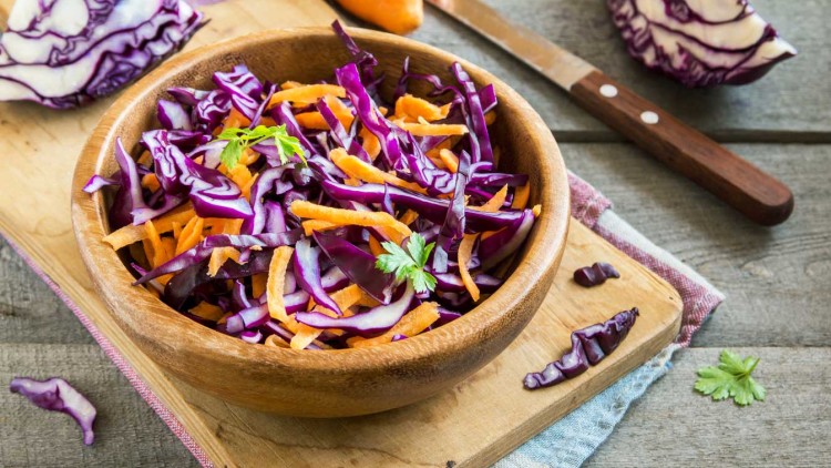 Holzschüssel mit Rotkohl- und Karottenstreifen, daneben Messer, Karotte und Rotkohlstücke auf Holzbrett.