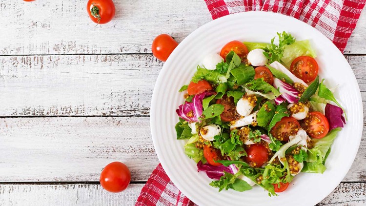 Ein Teller mit gemischtem Salat, darunter Tomaten, Mozzarella, grüne Blätter und Senfsamen, auf rot-weiß kariertem Stoff.