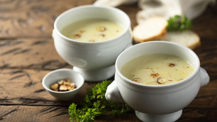 Zwei weiße Suppenschalen mit heller Cremesuppe und gehackten Nüssen, daneben eine kleine Schale Nüsse, Petersilie und Brot.
