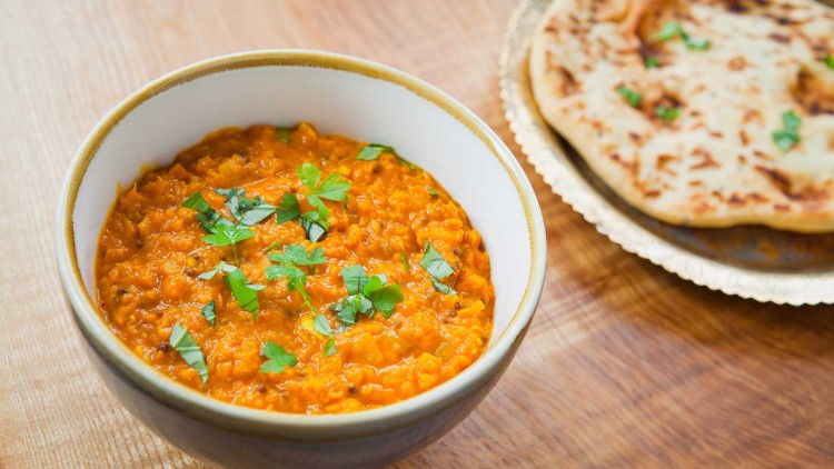 Eine weiße Schale mit orangem Linseneintopf, verziert mit grüner Petersilie, daneben ein Teller mit Naan-Brot auf Holzuntergrund.