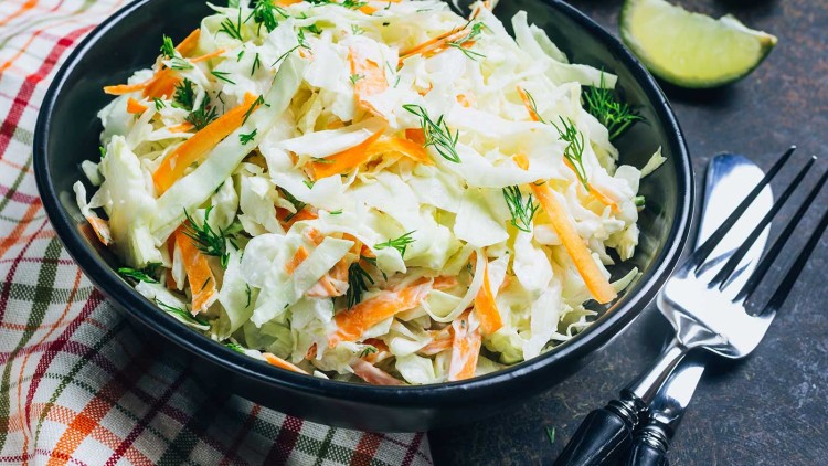 Schüssel mit Coleslaw aus Weißkohl, Möhren und Dill auf einem bunt karierten Tuch, daneben eine Gabel und Limettenspalten.