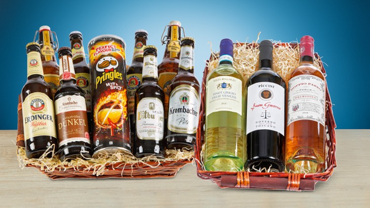 Zwei Geschenkkörbe mit verschiedenen Bierflaschen, einer Chipsdose und drei Weinflaschen auf hellem Holztisch vor blauem Hintergrund.