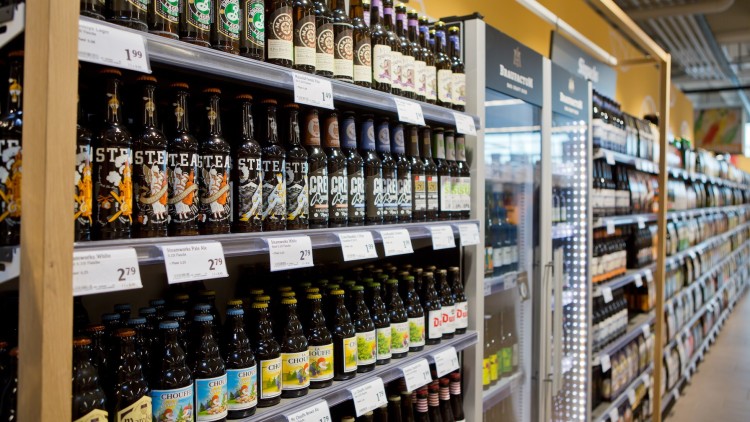 Regale mit verschiedenen Bierflaschen, Etiketten sichtbar, darüber Preisschilder, daneben ein Kühlschrank mit Getränken.