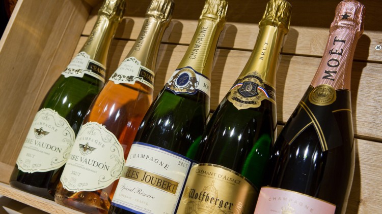 Sechs Champagner- und Cremantflaschen in unterschiedlichen Designs in einem hölzernen Regal nebeneinander platziert.