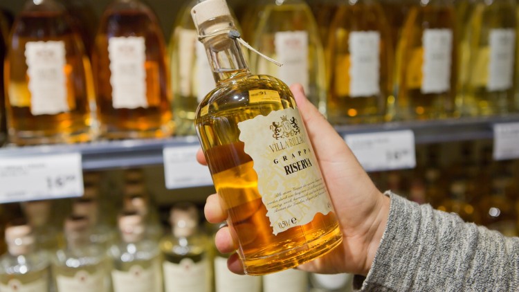 Hand hält eine Flasche „Villarzale Grappa Riserva“ vor einem Regal mit weiteren Spirituosenflaschen.