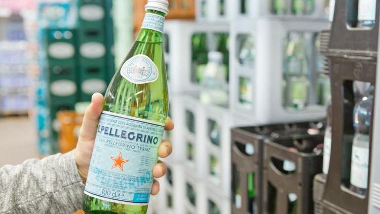 Eine Hand hält eine grüne Glasflasche S. Pellegrino vor einem Regal mit weiteren Getränkekisten.