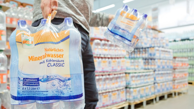 Person trägt einen 6er-Pack „GLOBUS Natürliches Mineralwasser mit Kohlensäure Classic“ durch einen Getränkemarkt.