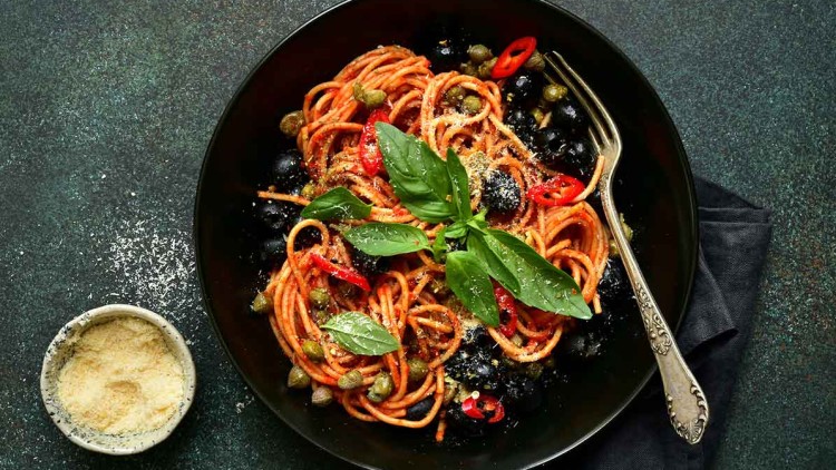 Schwarzer Teller mit Spaghetti, Tomatensauce, Oliven, Kapern, Chilischeiben, Basilikum und geriebenem Käse, daneben kleine Schale Käse.