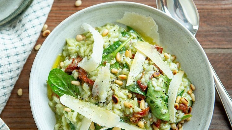 Cremiges Risotto mit Parmesan, Spinat, getrockneten Tomaten und Pinienkernen in einer hellen Schüssel auf Holztisch.